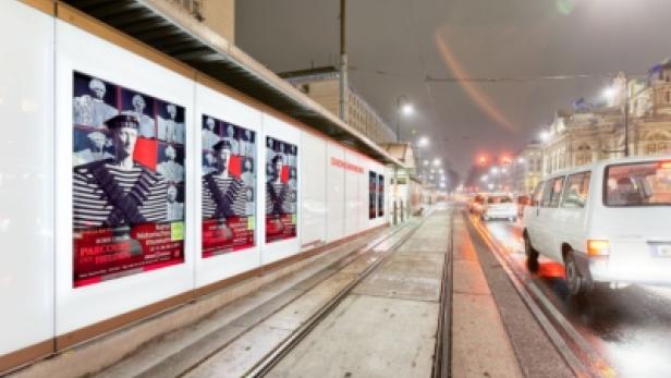 Eine Straßenbahnhaltestelle mit Plakaten und regennasser Fahrbahn bei Nacht.
