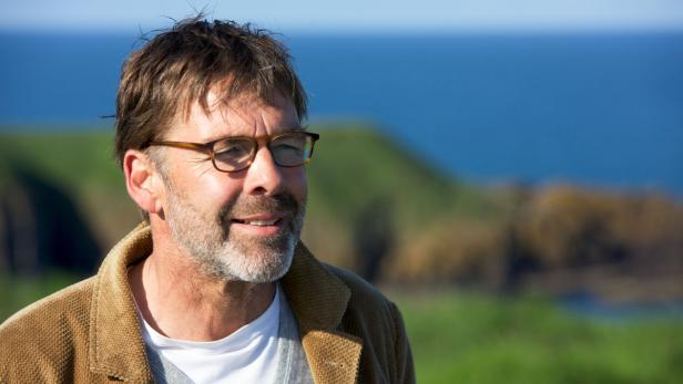 Ein Mann mit Brille und Bart blickt vor einer Küstenlandschaft in die Ferne.