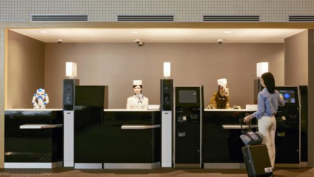 Eine Frau checkt in einem Hotel ein, dessen Rezeption von einem Roboter, einer menschlichen Angestellten und einem Dinosaurier besetzt ist.