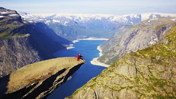 Eine Person sitzt auf dem Trolltunga-Felsen mit Blick auf einen norwegischen Fjord.