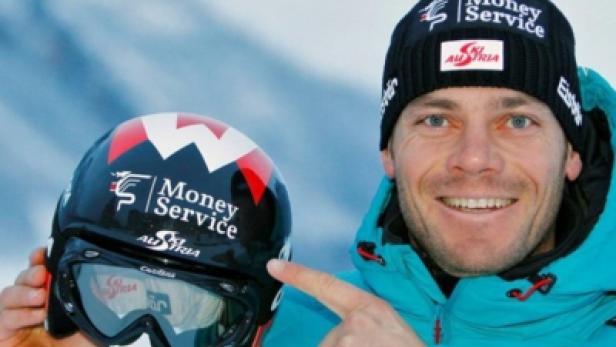 Ein Mann mit Mütze zeigt auf einen Skihelm mit der Aufschrift „Money Service Austria“.