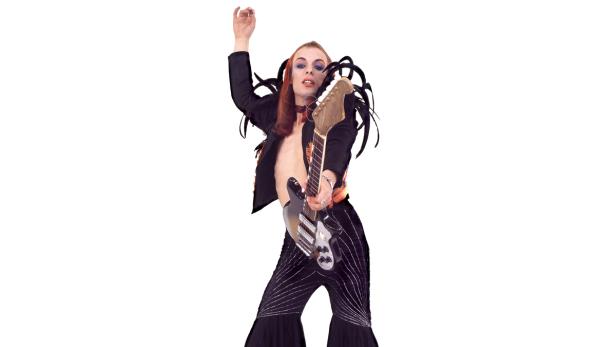 Ein androgyn wirkender Musiker mit Gitarre in einem extravaganten Outfit.