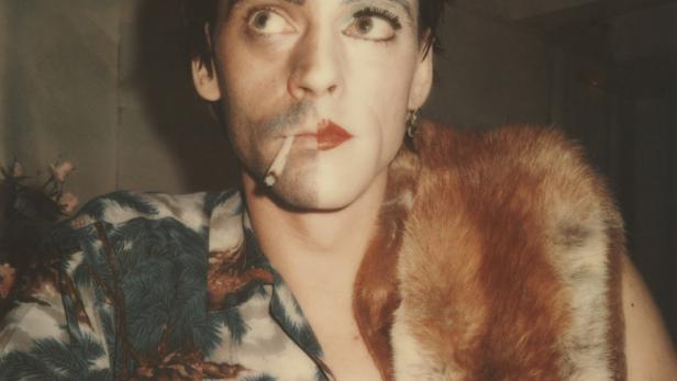 Ein Mann mit Make-up und einem Fuchsfell über der Schulter raucht eine Zigarette.