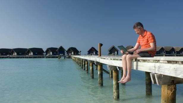 Ein Mann sitzt barfuß auf einem Holzsteg am Meer und arbeitet am Laptop.
