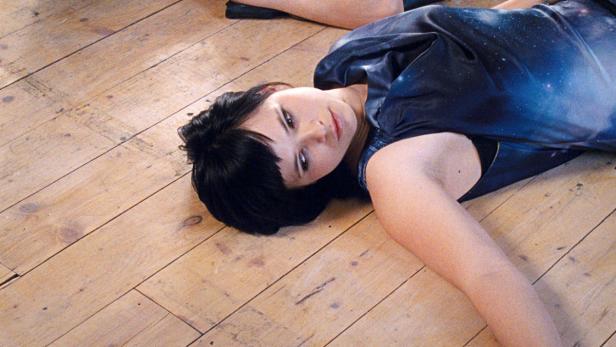 Eine Frau mit dunklen Haaren liegt auf einem Holzboden und trägt ein Kleid mit Weltraummuster.
