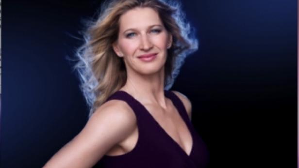 Eine Frau mit blonden Haaren und einem violetten Kleid posiert vor einem dunklen Hintergrund.