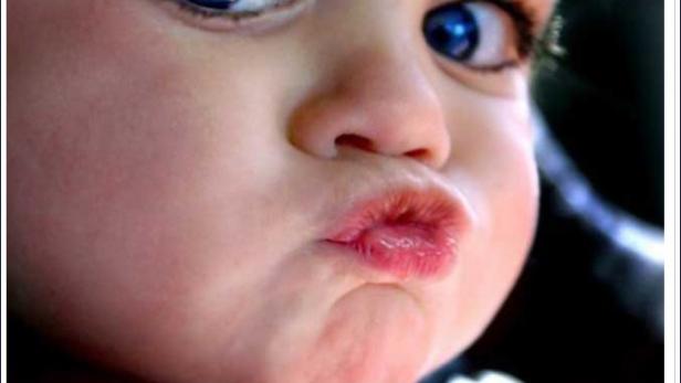 Ein Baby mit großen blauen Augen schaut in die Kamera und spitzt die Lippen.