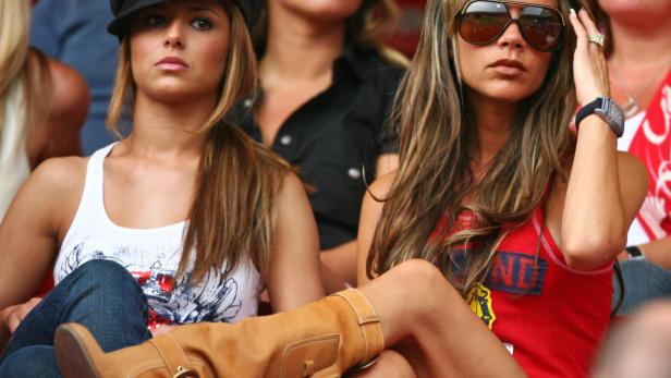 Cheryl Cole mit Stiefeln und Victoria Beckham mit Sonnenbrille sitzen auf der Tribüne.