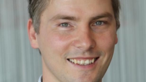 Ein lächelnder Mann in Anzug und Hemd vor einem hellen Hintergrund.