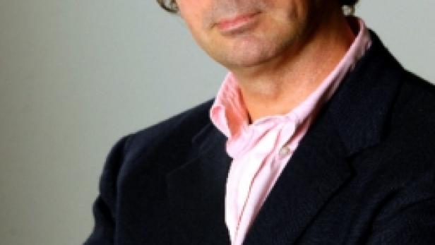 Ein Mann mit Brille und verschränkten Armen in einem dunklen Anzug und rosa Hemd.