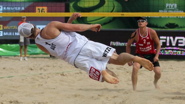 Ein Beachvolleyballspieler (AUT) springt, um den Ball zu erreichen, während ein Spieler (POL) zusieht.
