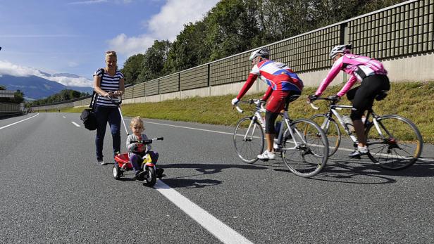 Eine Mutter zieht ihr Kind auf einem Dreirad, während zwei Radfahrer auf einer Straße fahren.