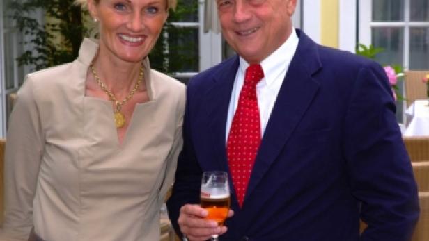 Ein Mann im Anzug und eine Frau in einem beigen Kleid posieren lächelnd für ein Foto.