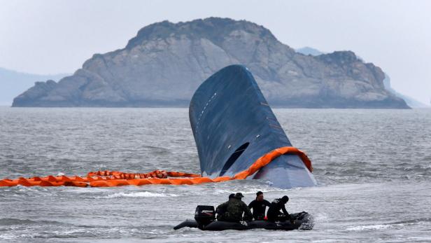Ein gekentertes Schiff liegt im Wasser, umgeben von einer orangefarbenen Ölsperre, während ein Rettungsboot sich nähert.