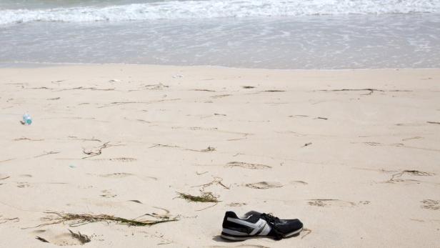 Ein einzelner Sportschuh liegt im Sand am Strand, im Hintergrund das Meer.
