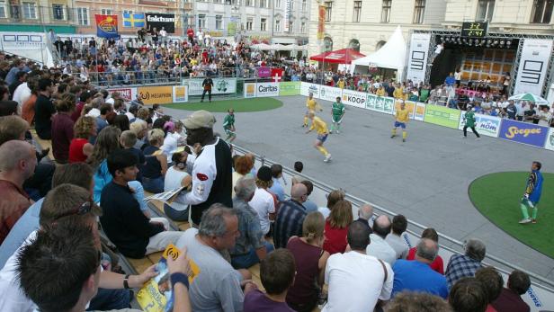 Zuschauer beobachten ein Straßenfußballspiel auf einem Platz mit Bühne.