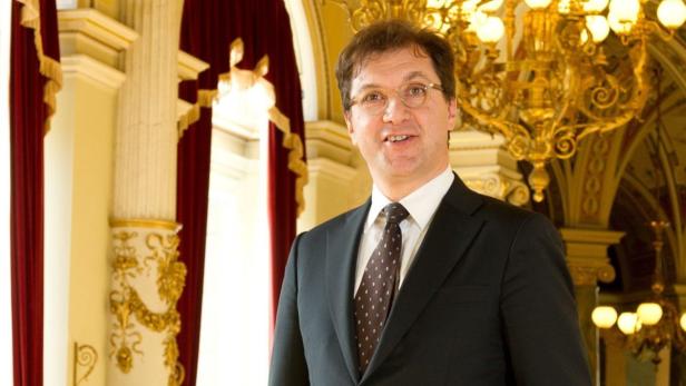 Ein Mann in Anzug und Krawatte steht in einem eleganten Saal mit goldenem Kronleuchter.