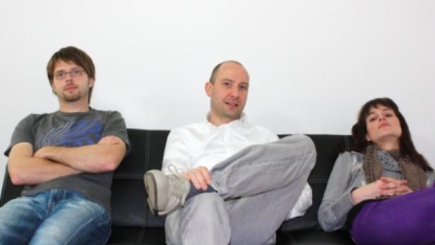 Drei Personen sitzen auf einer schwarzen Couch vor einer weißen Wand.