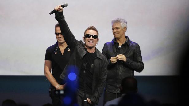 Bono von U2 mit erhobenem Mikrofon auf einer Bühne, flankiert von zwei weiteren Männern.