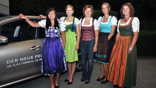 Fünf junge Frauen in Dirndln posieren neben einem Auto.