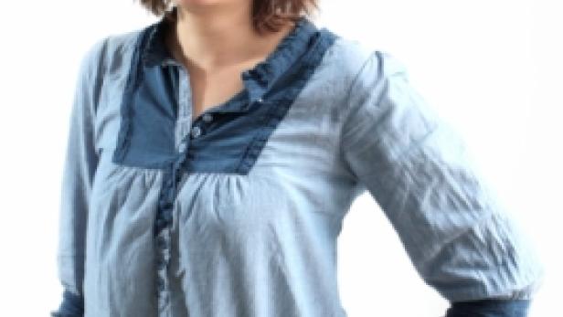 Eine lächelnde Frau mit braunen Haaren und einer hellblauen Bluse posiert vor einem weißen Hintergrund.
