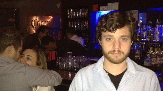 Ein Mann steht in einer Bar, im Hintergrund ein Paar beim Küssen.