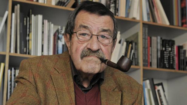 Ein älterer Mann mit Brille und Schnurrbart raucht eine Pfeife vor einem Bücherregal.