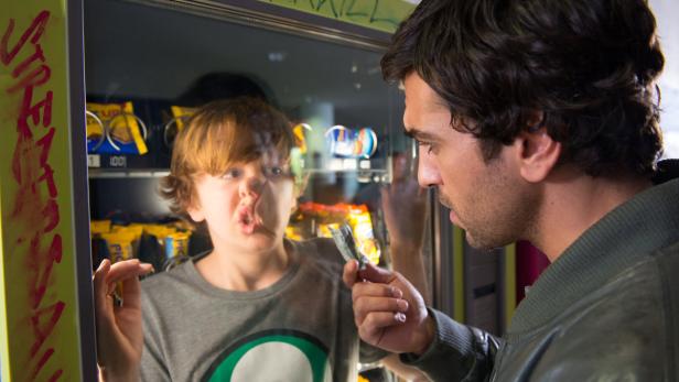 Ein Mann hält Geld vor einen Snackautomaten, in dem sich ein Kind spiegelt.