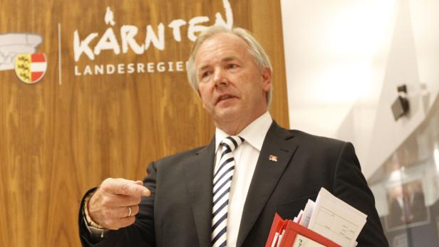 Ein Mann im Anzug gestikuliert vor einem Schild der Kärntner Landesregierung.