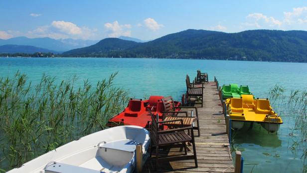 Ein Steg mit Tretbooten und Stühlen führt in einen ruhigen See vor einer bewaldeten Hügellandschaft.