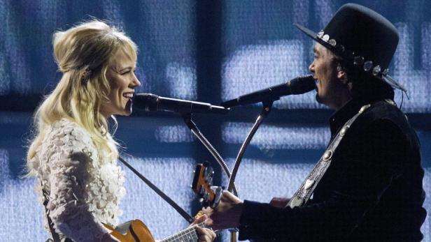 Eine blonde Frau und ein Mann mit Hut singen und spielen Gitarre auf einer Bühne.