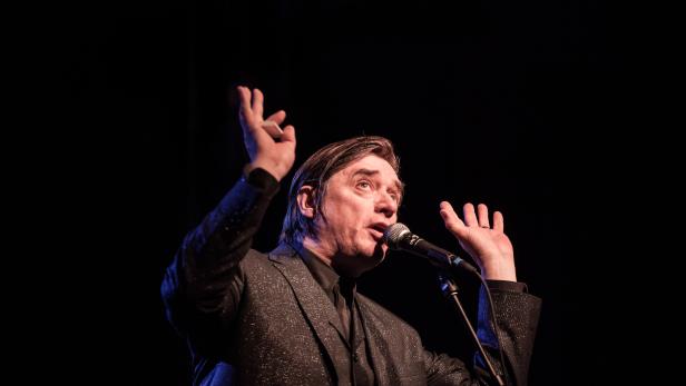 Ein Mann in einem glitzernden Anzug singt mit erhobenen Händen in ein Mikrofon.