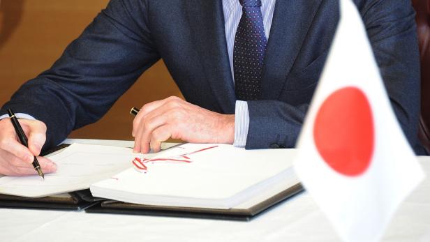 Ein Mann im Anzug unterzeichnet ein Dokument neben einer japanischen Flagge.