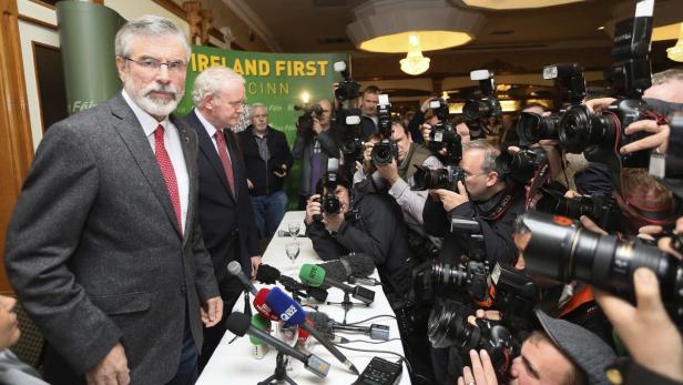 Gerry Adams und ein weiterer Mann werden von einer Gruppe Fotografen umringt.