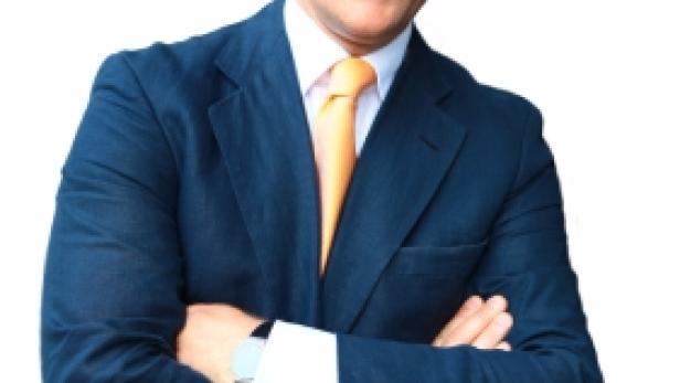 Ein Mann im Anzug mit verschränkten Armen lächelt.