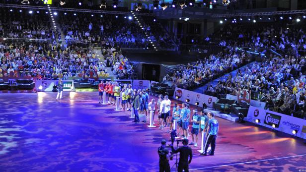 Eine Halle mit Zuschauern und zwei Teams, die sich auf einem Spielfeld gegenüberstehen.