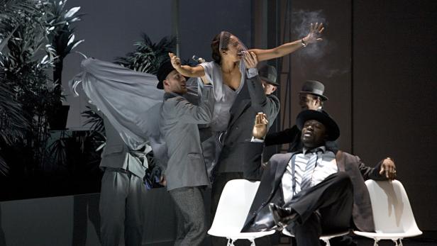 Eine Theaterszene mit einer Frau, die von mehreren Männern in Anzügen umgeben ist.