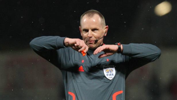 Ein Fußballschiedsrichter mit erhobenen Händen und einer Pfeife im Mund.