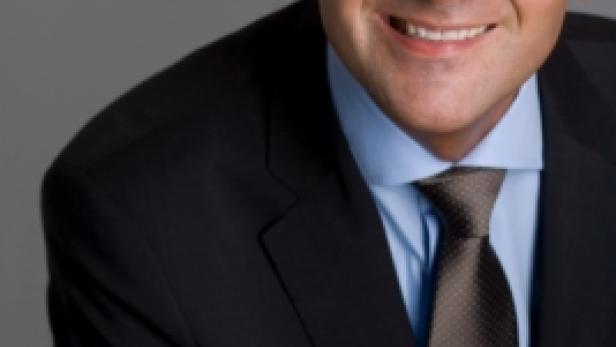 Ein lächelnder Mann in Anzug und Krawatte vor einem grauen Hintergrund.
