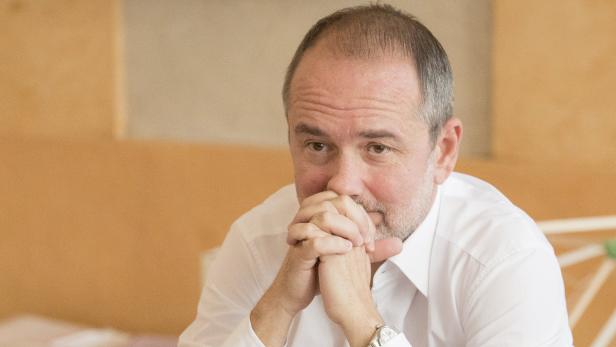 Ein Mann in einem weißen Hemd sitzt mit verschränkten Händen vor dem Gesicht.