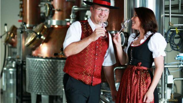 Ein Mann und eine Frau in Tracht stehen vor einer Destillieranlage und halten Weingläser.