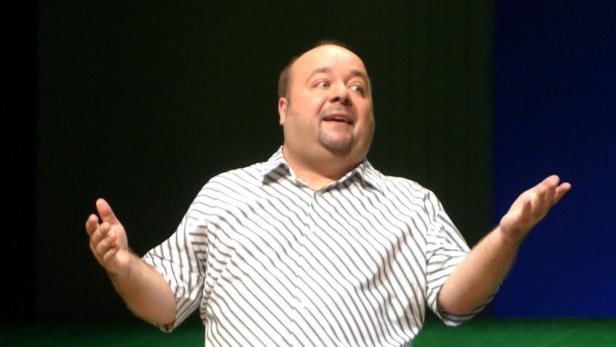 Ein Mann mit gestreiftem Hemd gestikuliert vor einem schwarzen und blauen Hintergrund.