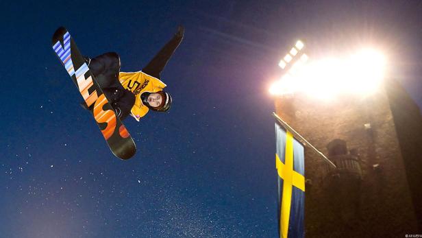 Eine Snowboarderin springt vor einer schwedischen Flagge und Flutlicht in der Luft.
