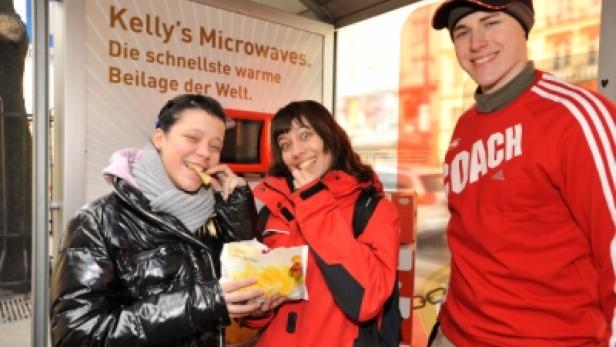 Drei Personen essen Kelly's Microwaves Chips vor einem Werbeplakat.