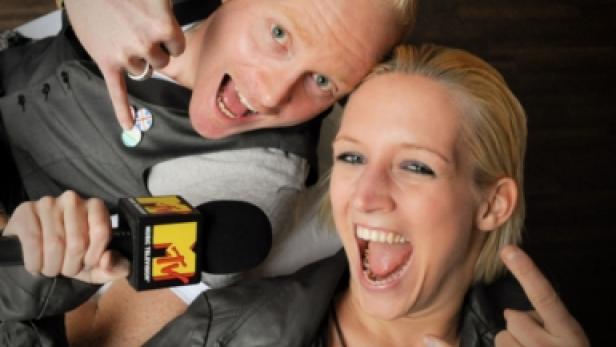 Zwei blonde Personen posieren mit einem MTV-Mikrofon und machen Handzeichen.