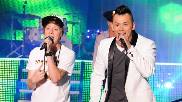 Zwei Sänger treten auf einer Bühne mit Mikrofonen vor einem farbigen Hintergrund auf.