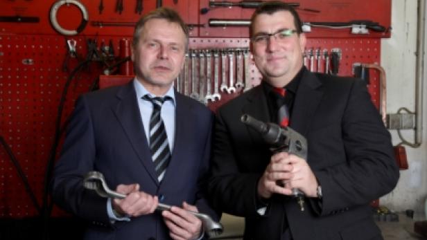 Zwei Männer in Anzügen halten Werkzeuge vor einer Werkzeugwand.