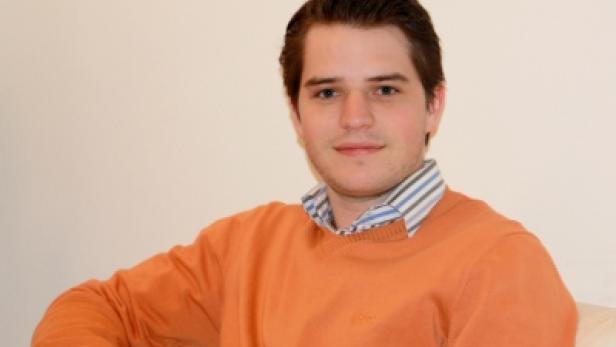 Ein junger Mann mit einem orangefarbenen Pullover sitzt auf einem Sofa.