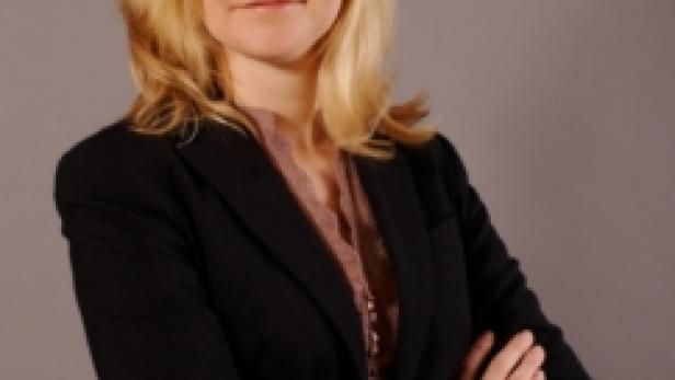 Eine blonde Frau mit verschränkten Armen vor einem grauen Hintergrund.
