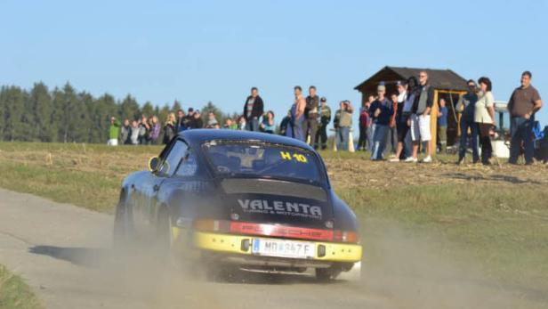 Ein schwarzer Porsche fährt auf einer staubigen Straße an einer Zuschauermenge vorbei.